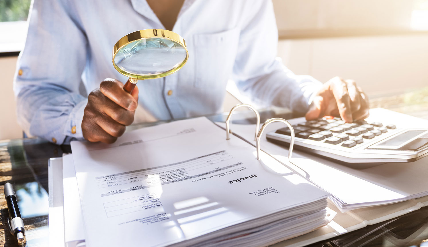 office worker examining folder of paperwork with magnifying glass, using calculator in other hand